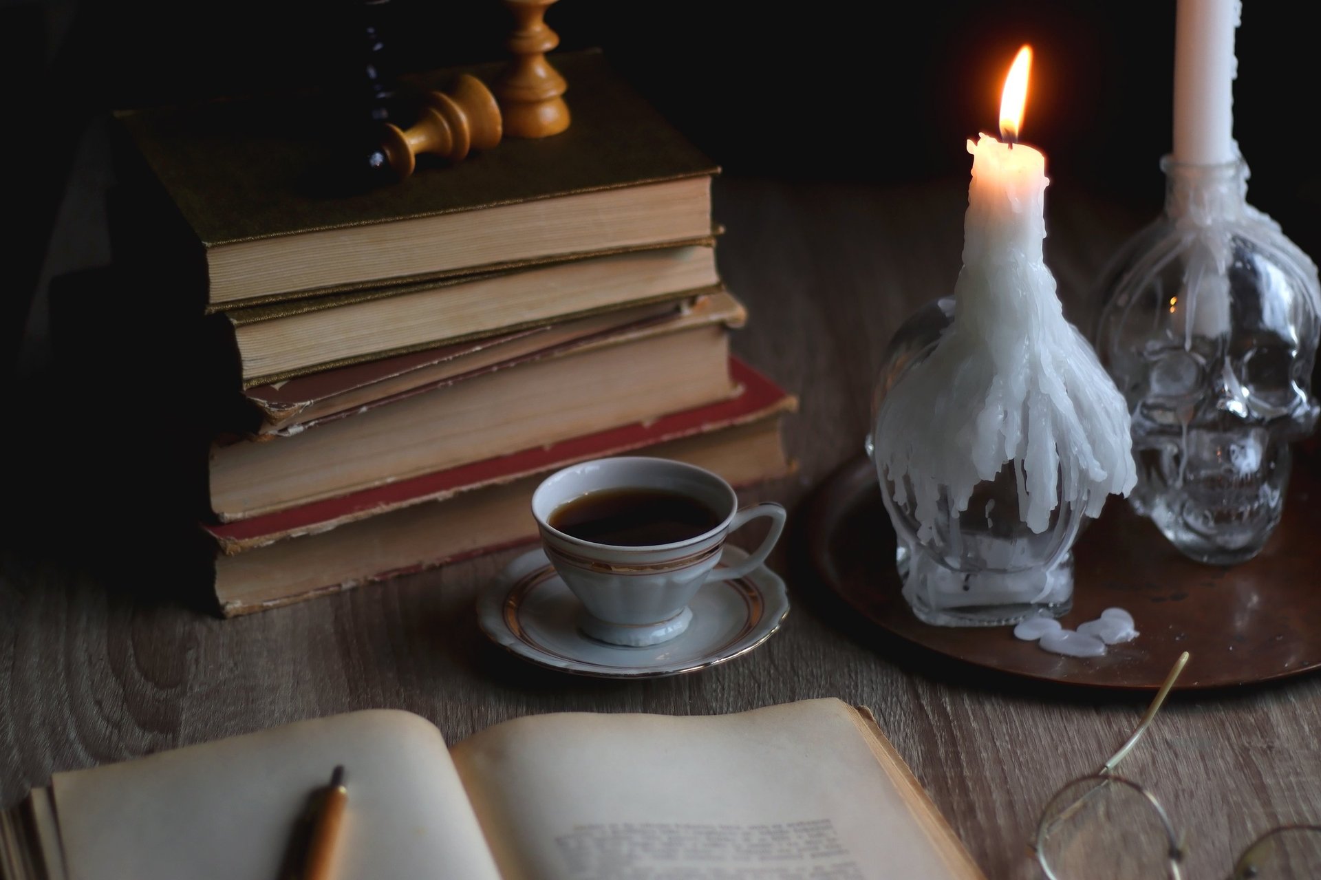 Stack of vintage books, cup of tea or coffee, lit candles, reading glasses and chess pieces on wooden table