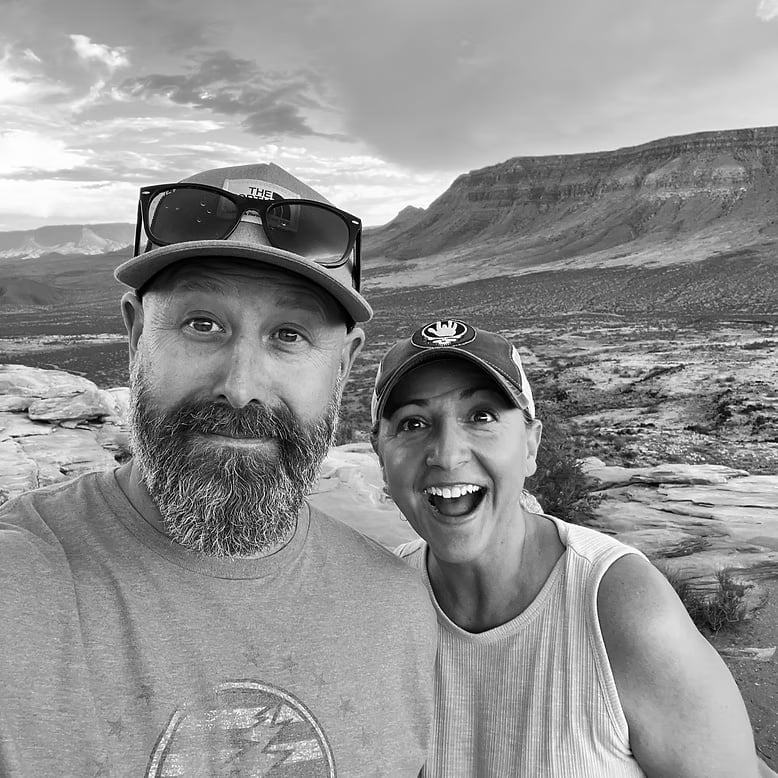 Black and white photo of a bearded man and smiling woman wearing caps in a desert mountain landscape